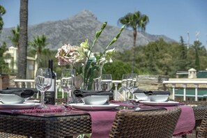 A table set for dining with a bottle of wine, glasses, and a vase of flowers, overlooking a mountainous landscape. - Nueva Andalucia Penthouse, Golf View, Pool (Marbella)