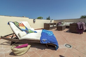 Ein Liegestuhl mit Kissen, ein gewebter Korb und ein blaues Handtuch auf einer gefliesten Terrasse.