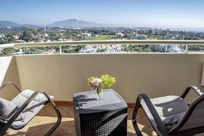 A balcony with two chairs, a small table, and a vase with flowers. A cityscape with mountains is visible in the background. - LL- 1 bedroom apt close to Laguna Village Estepona (Estepona)