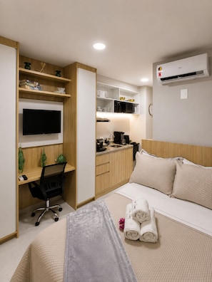 Ein kleines Hotelzimmer mit einem Bett, einem Schreibtisch mit Stuhl, einem Fernseher, einer Küchenzeile und einer Klimaanlage.
