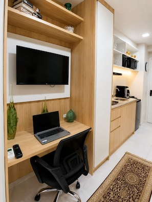 Espaço compacto com televisão de tela plana acima de uma escrivaninha de madeira, um laptop, uma cadeira de escritório preta e uma pequena cozinha com micro-ondas e máquina de café.