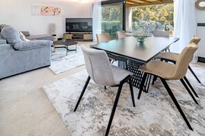 Una sala moderna con un sofá gris, una mesa de comedor con sillas, un televisor y un ventanal con vistas a árboles.
