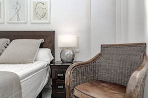Ein Schlafzimmer mit einem Bett, einem Nachttisch mit Lampe, einem Korbstuhl und Kunstwerken an der Wand.