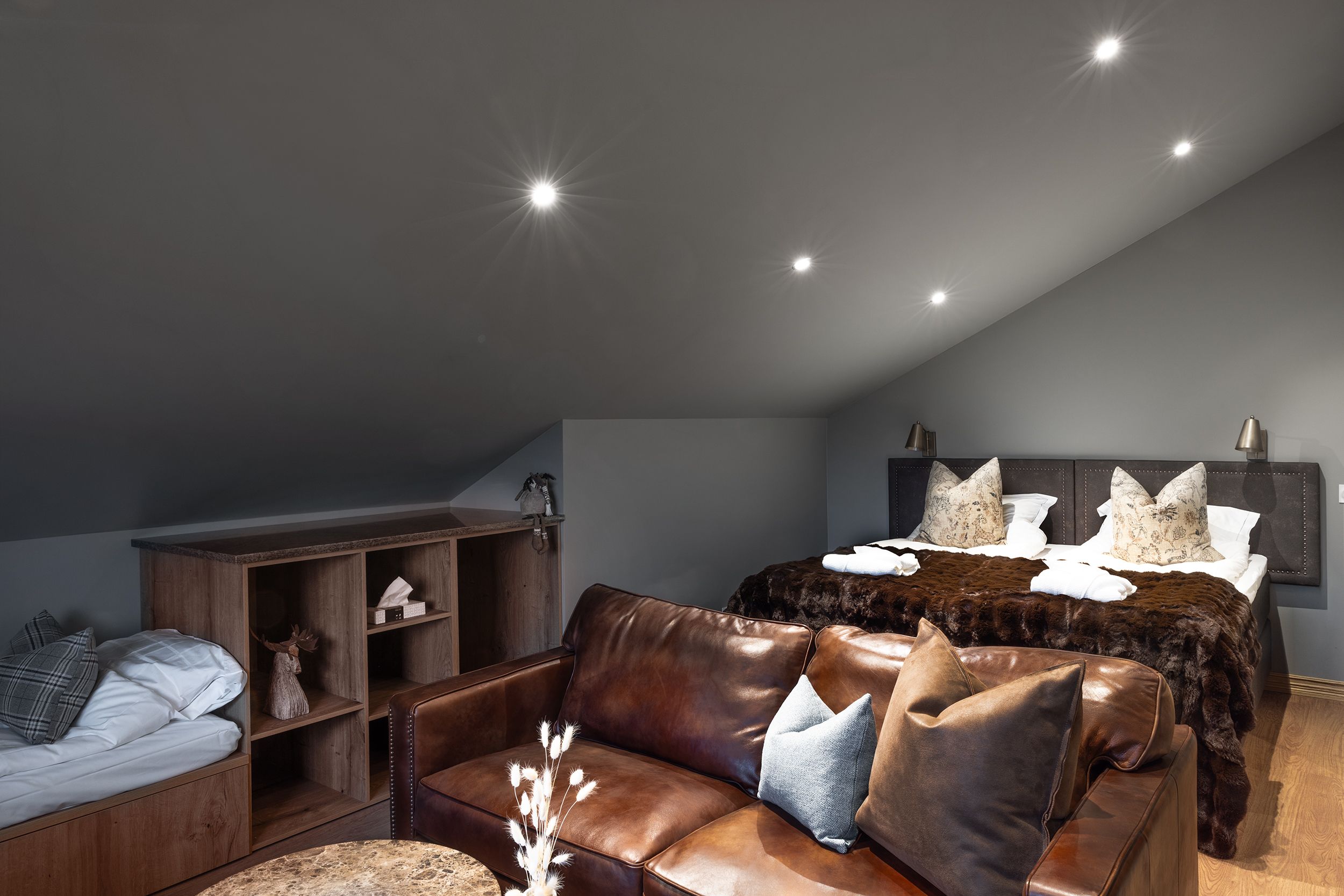 A modern bedroom with a sloped ceiling, a large bed with decorative pillows, a leather sofa, and a wooden cabinet.
