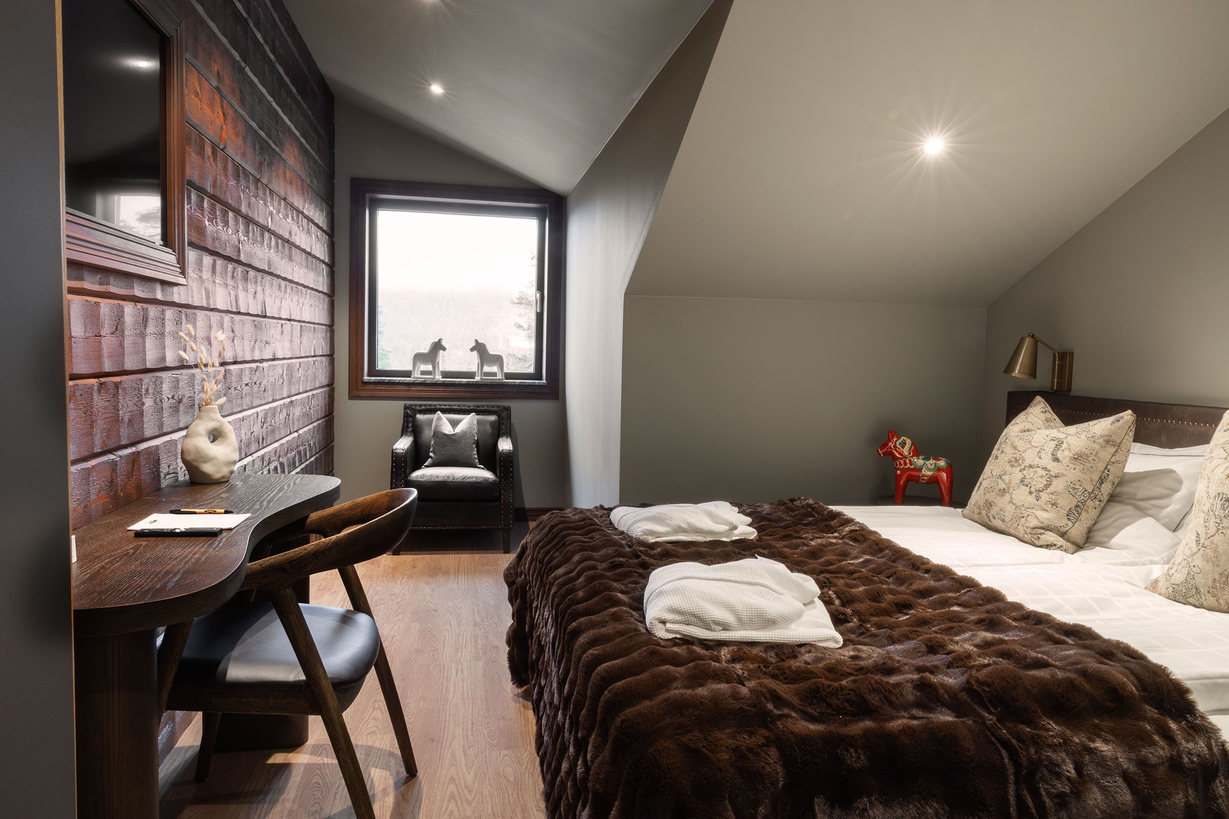 A bedroom with a large window, a desk, a chair, a bed with a fur throw, and a nightstand with a lamp.
