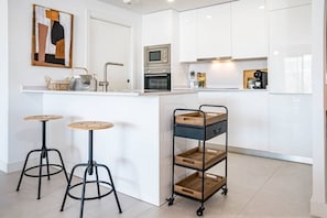 A modern kitchen with white cabinetry, a built-in oven, a microwave, and a coffee machine. There are two stools with round wooden tops and black metal legs. A wooden cart with drawers is also visible. - LMe4BB Sb4bb- apotel las mesas by roomservices (Estepona)