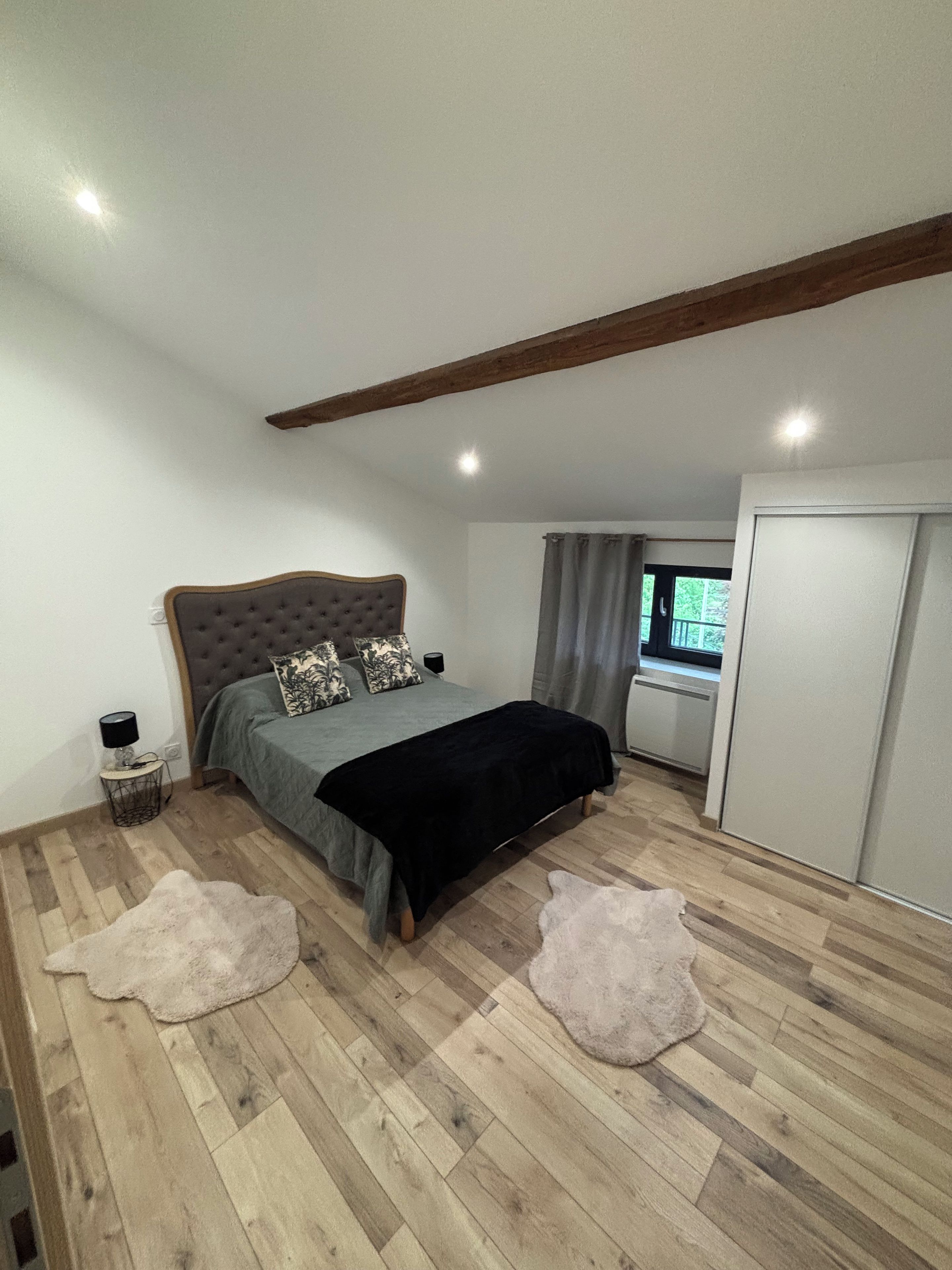 A bedroom with a bed, a wardrobe, a window with curtains, and a wooden ceiling beam.