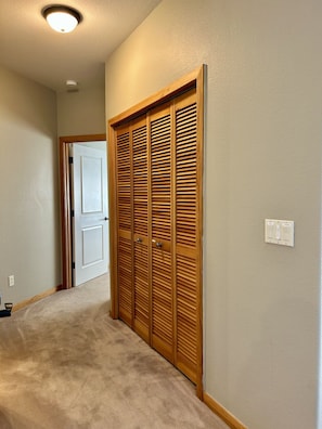Um corredor com piso coberto por carpete, um guarda-roupa de madeira com portas venezianas, uma porta branca ao fundo e um interruptor de luz na parede.