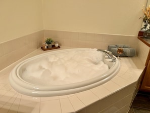 Ein Badezimmer mit einer Badewanne voller Blasen, einem Holzregal mit Pflegeprodukten und einem Spiegel an der Wand.