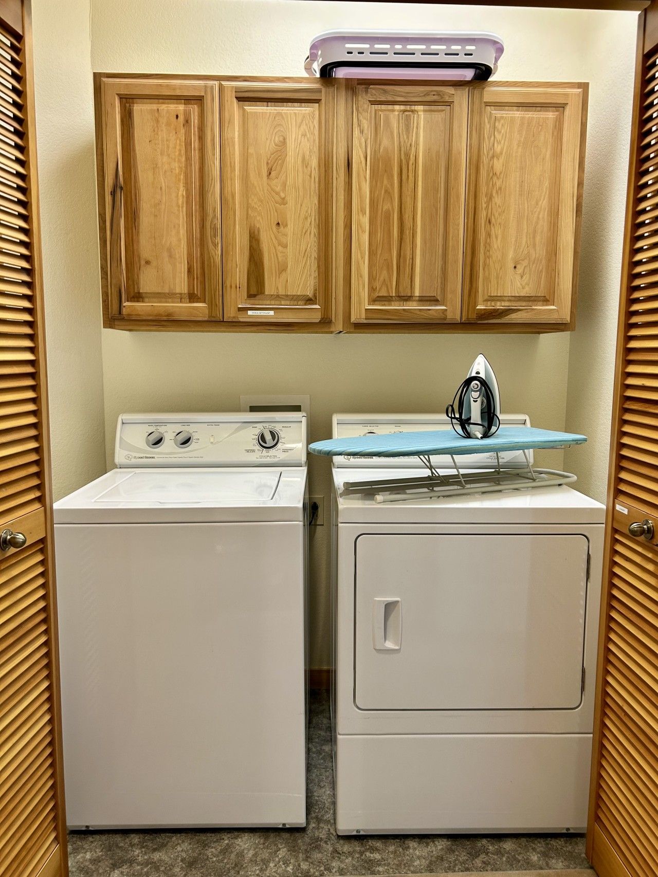 Un espace buanderie équipé de deux machines à laver et d’un sèche-linge, avec des placards en bois au-dessus et une petite planche à repasser.