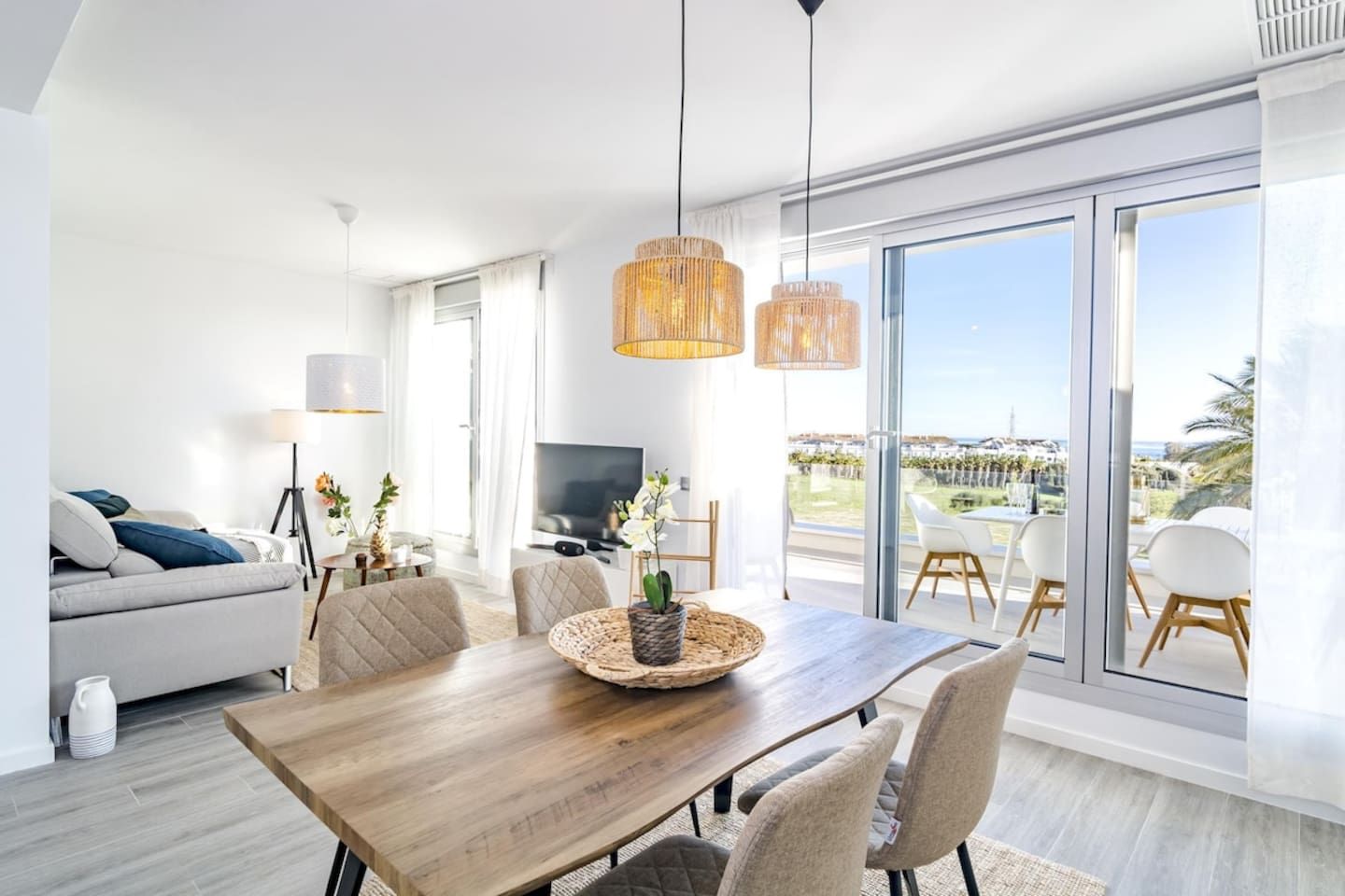 A modern living room with a wooden dining table, comfortable seating, a television, and large windows offering a view of the outdoors.