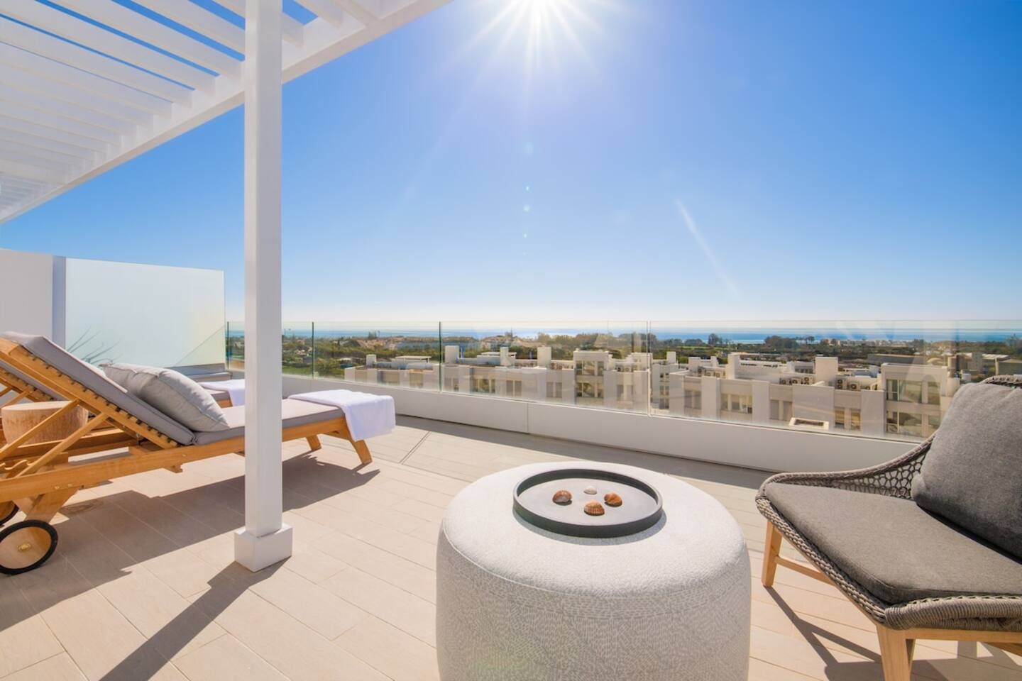 A rooftop terrace with a clear view of the cityscape, a lounge chair, a small round table with decorative items, and a white structure.