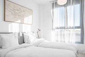 Quarto com cama, mesa de cabeceira, luminária e janela com cortinas finas.