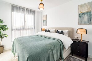 Ein Schlafzimmer mit einem Bett, Nachttischlampen, einem Fenster mit durchsichtigen Vorhängen, einer Topfpflanze und Bildern an den Wänden.