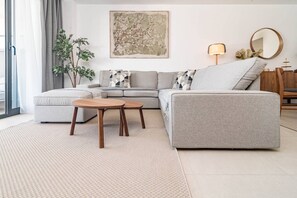 Uma sala de estar moderna com um sofá cinzento, duas mesas laterais de madeira, um quadro abstrato grande, um abajur e um espelho na parede.
