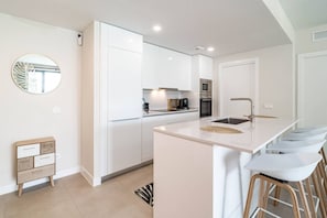 A modern kitchen with white cabinetry, a central island with bar stools, and a round mirror on the wall. - LMe2BA- Cozy ground floor flat in luxury resort (Estepona)