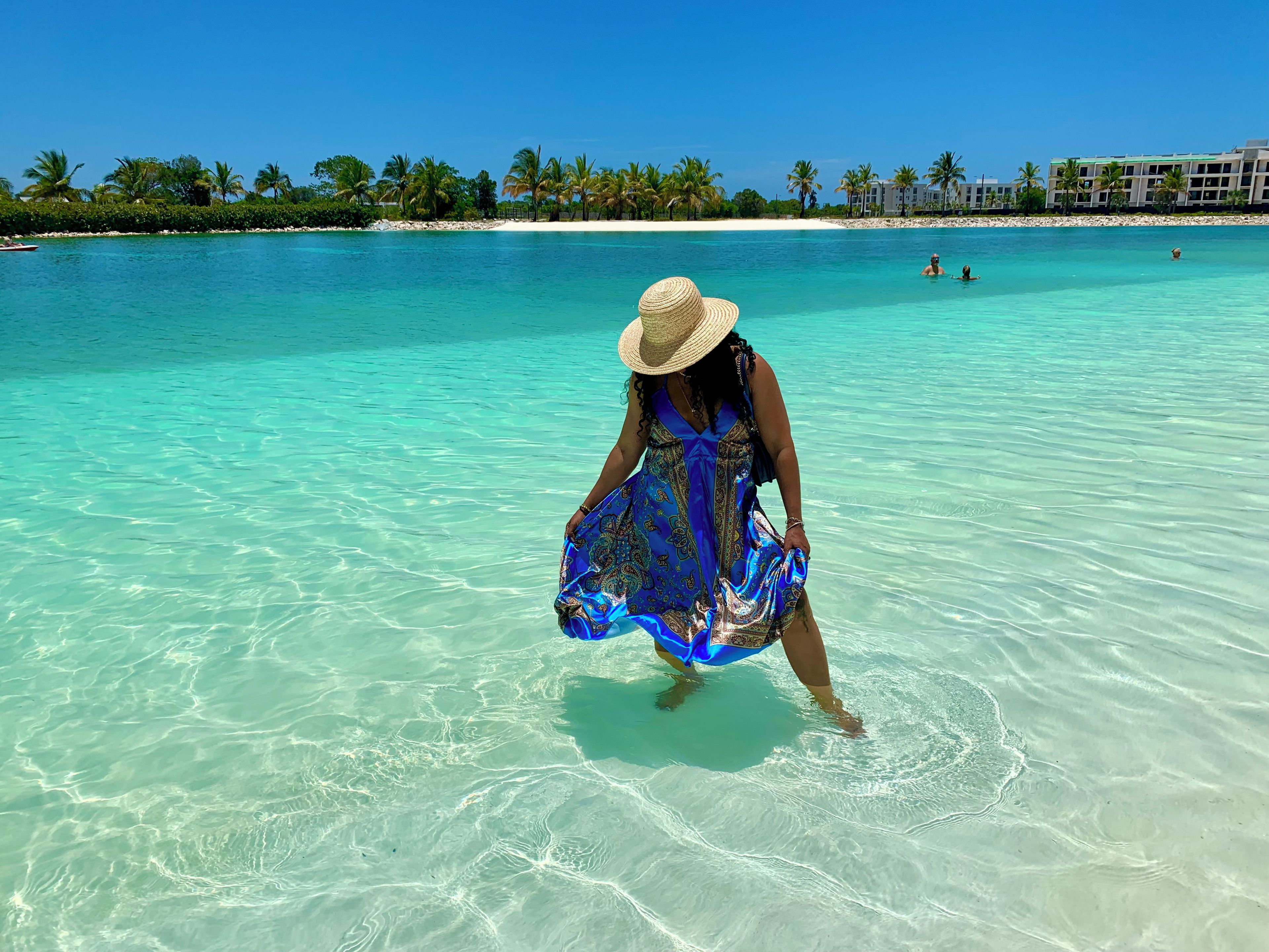Eine Frau im blauen Kleid und Strohhut geht durch klares türkisblaues Wasser, im Hintergrund ein Strand und Palmen.