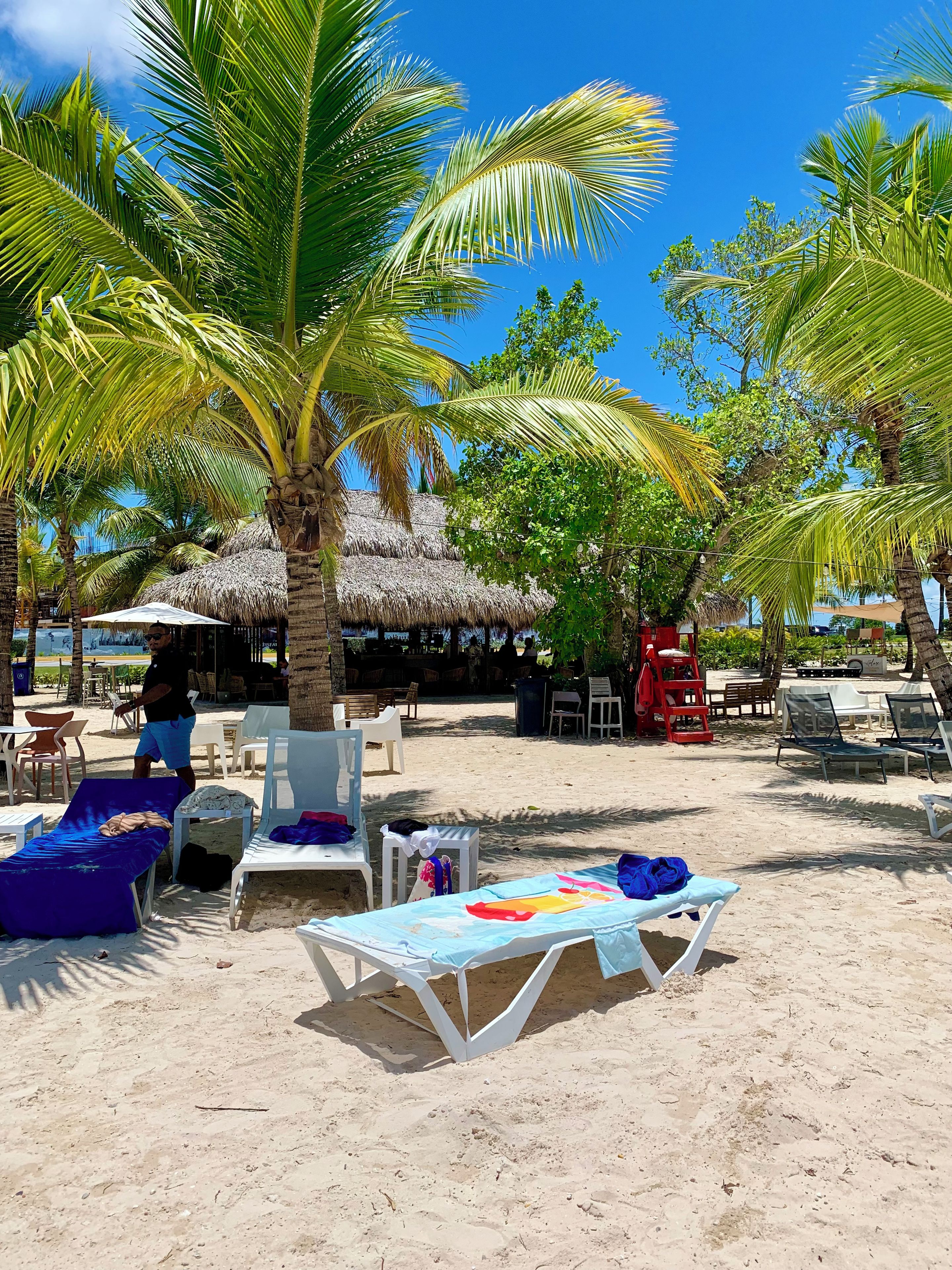 Un espace de détente aménagé au bord de la plage, avec des structures à toits de chaume, des chaises longues blanches et un sol sablonneux.