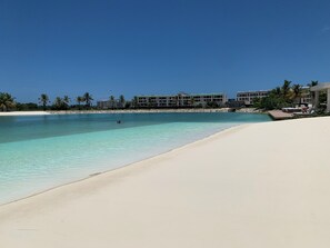 Un resort en la playa con un mar azul cristalino, una orilla arenosa y edificios del resort al fondo.