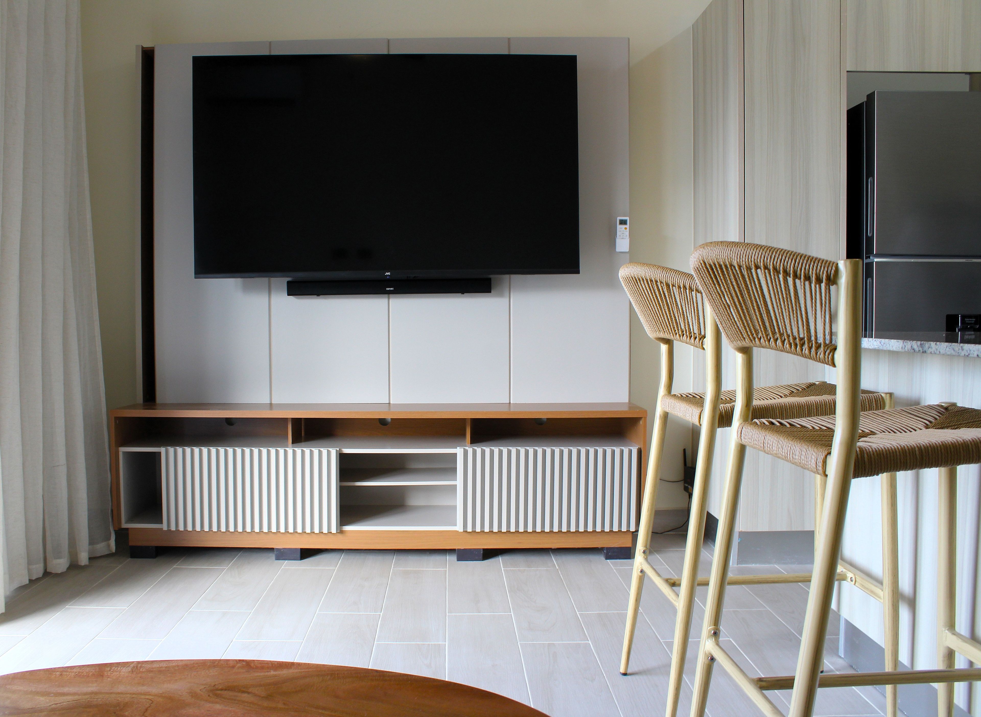 A modern living room with a flat-screen TV mounted on the wall, a wooden entertainment unit below, and a pair of wicker bar stools.
