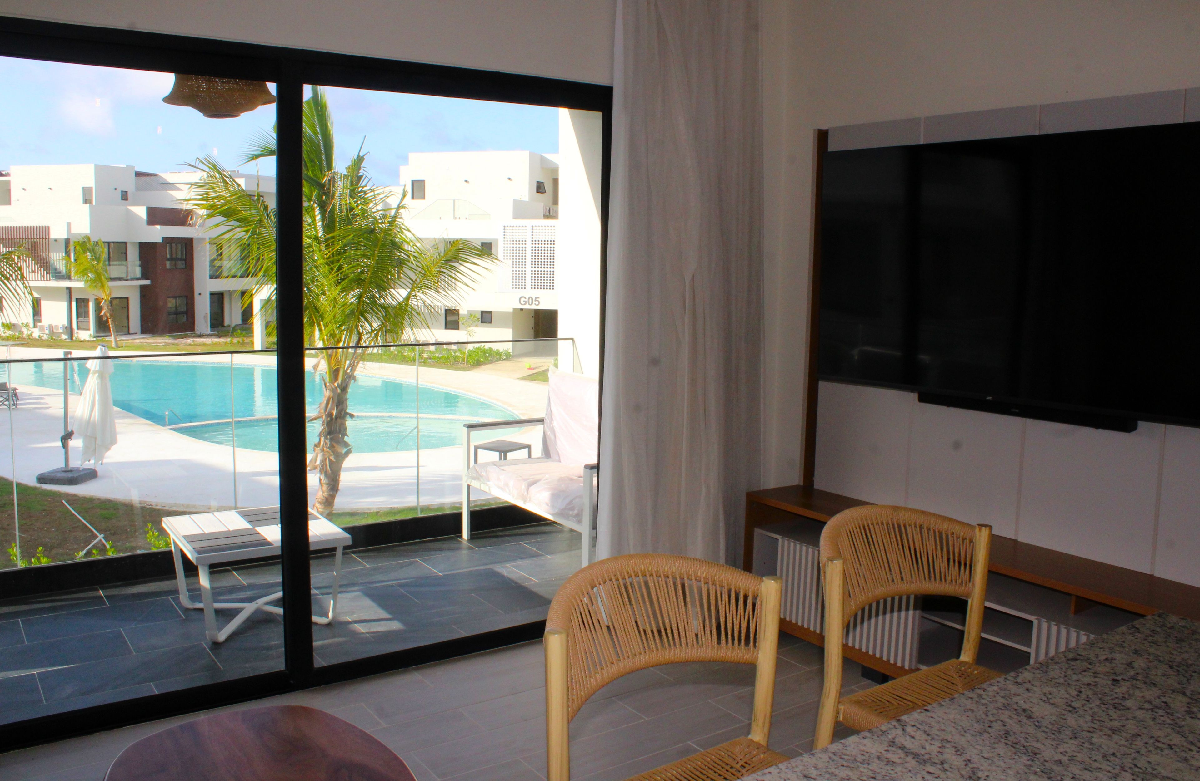 Quarto moderno com uma janela ampla de frente para uma piscina e um edifício identificado como G05. Há uma cadeira de vime e uma mesa pequena no quarto.