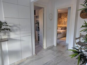 A hallway with a white tiled wall, a door leading to a room with a patterned wall, and a plant on the right. - Sea-View Apartment, Walk to Beach, Estepona (Estepona)