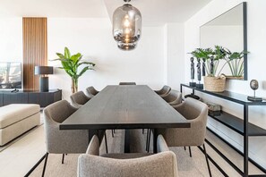 Ein moderner Essraum mit einem langen Tisch, mehreren Stühlen, einer Pendelleuchte, einer Pflanze und einem Sideboard.