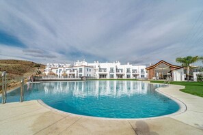 A swimming pool with a clear blue water surface, surrounded by a concrete deck. In the background, there are white buildings with multiple windows and balconies. - PA18- Stunning flat, gym, indoor and outdoor pool (La Alquería-El Paraíso)