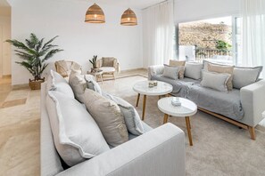 Een moderne woonkamer met een grijze bank, twee ronde witte tafels, rieten stoelen en een grote potplant.