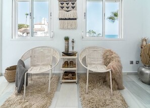 Ein Zimmer mit zwei Korbstühlen, einem Teppich, einer kleinen Ablagefläche mit Dekoration und Meerblick durch große Fenster.