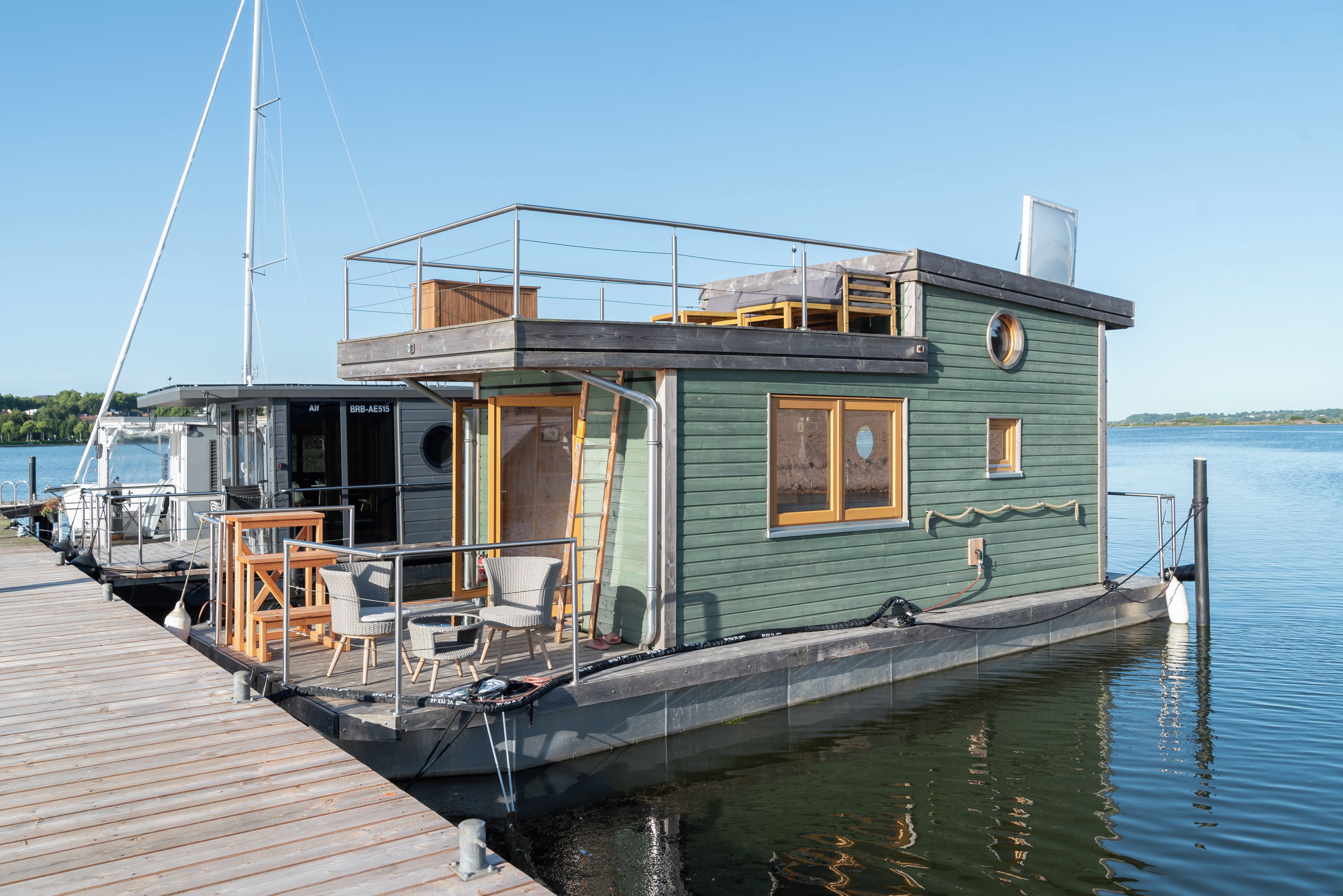 Ein Hausboot an einem Steg, mit einer Außenterrasse, Sitzgelegenheiten im Freien und einem kleinen Fenster.