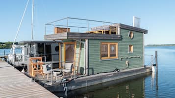 Ein Hausboot an einem Steg, mit einer Außenterrasse, Sitzgelegenheiten im Freien und einem kleinen Fenster.