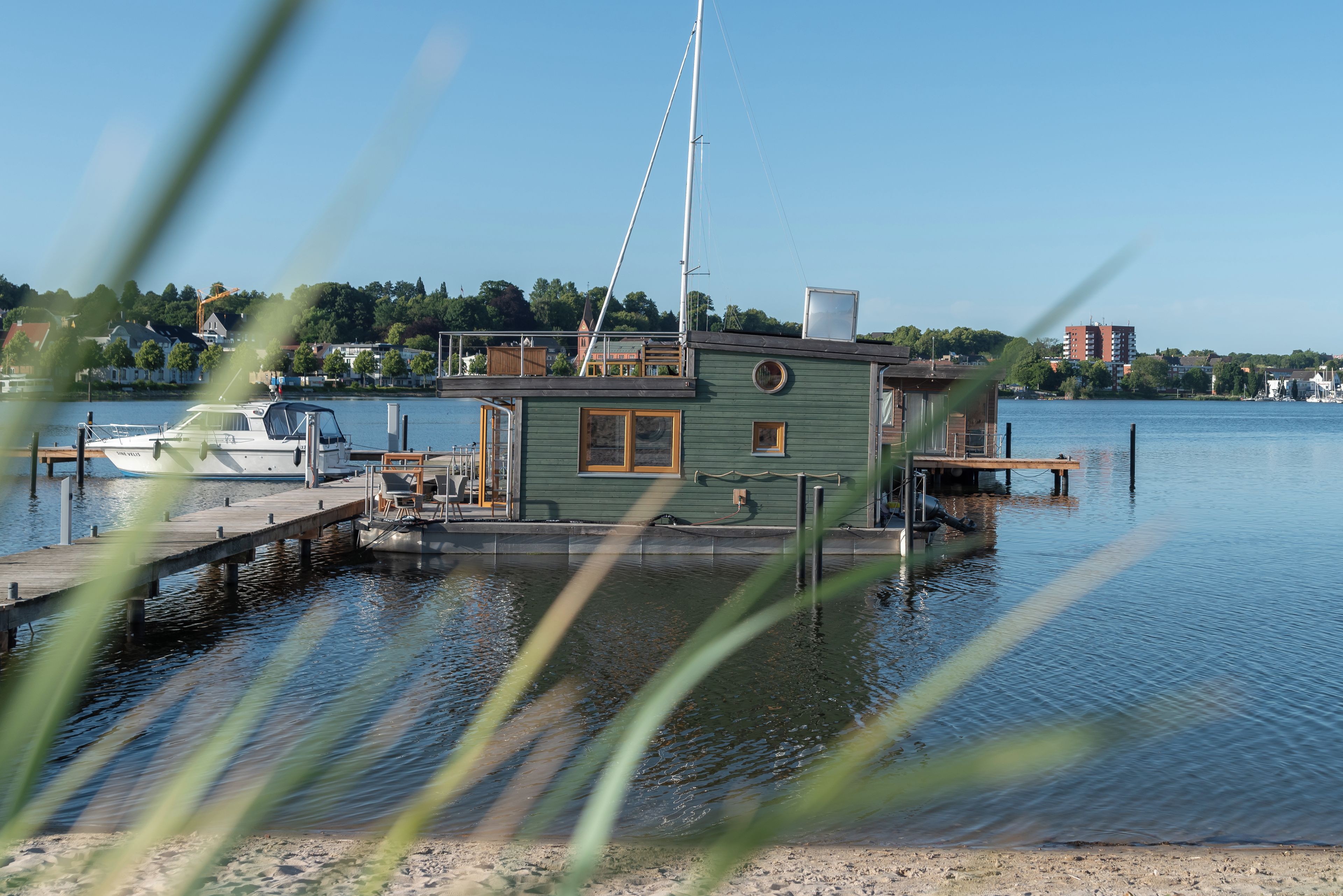 Ein Hausboot liegt an einem Steg, mit freiem Blick auf das Wasser und eine ferne Stadt im Hintergrund.