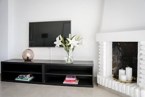 A modern living room with a flat-screen TV mounted on the wall, a black entertainment unit with books, a clear vase with white flowers, and a white fireplace with candles. - RDM - Stylish Holiday Apartment with Ocean Views (Mijas)
