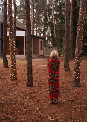 A person wrapped in a blanket standing in a forested area with trees and a building in the background. - Modern Barn with SPA, 5 km from Zegrze Reservoir (Beniaminów)