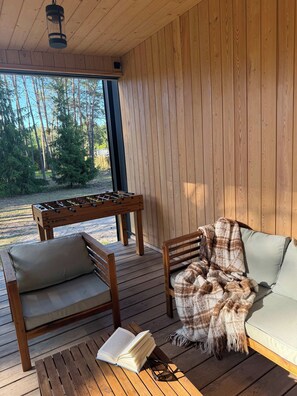Una terraza de madera con una mesa de fútbol de mesa, dos sillones acolchados, una manta y un libro abierto.