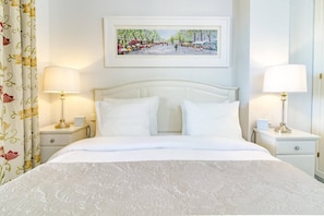 Habitación de hotel con una cama grande, cortinas blancas, dos lámparas de noche y un cuadro enmarcado en la pared.