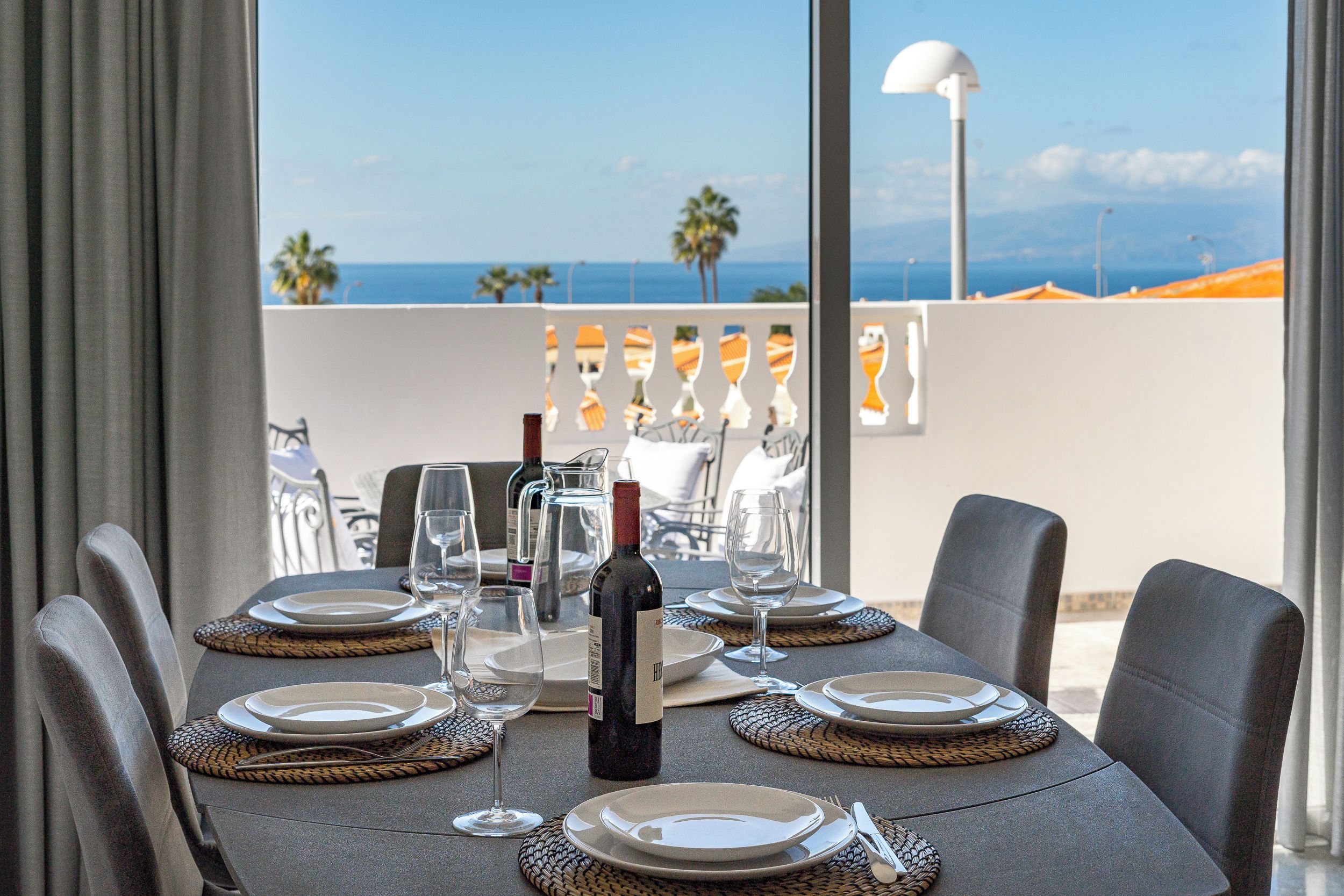 A table set for dining with a view of the sea, a bottle of wine, and a clear sky.