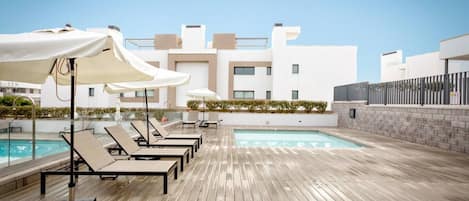 A rooftop pool area with lounge chairs and umbrellas, a clear sky, and a modern building in the background.