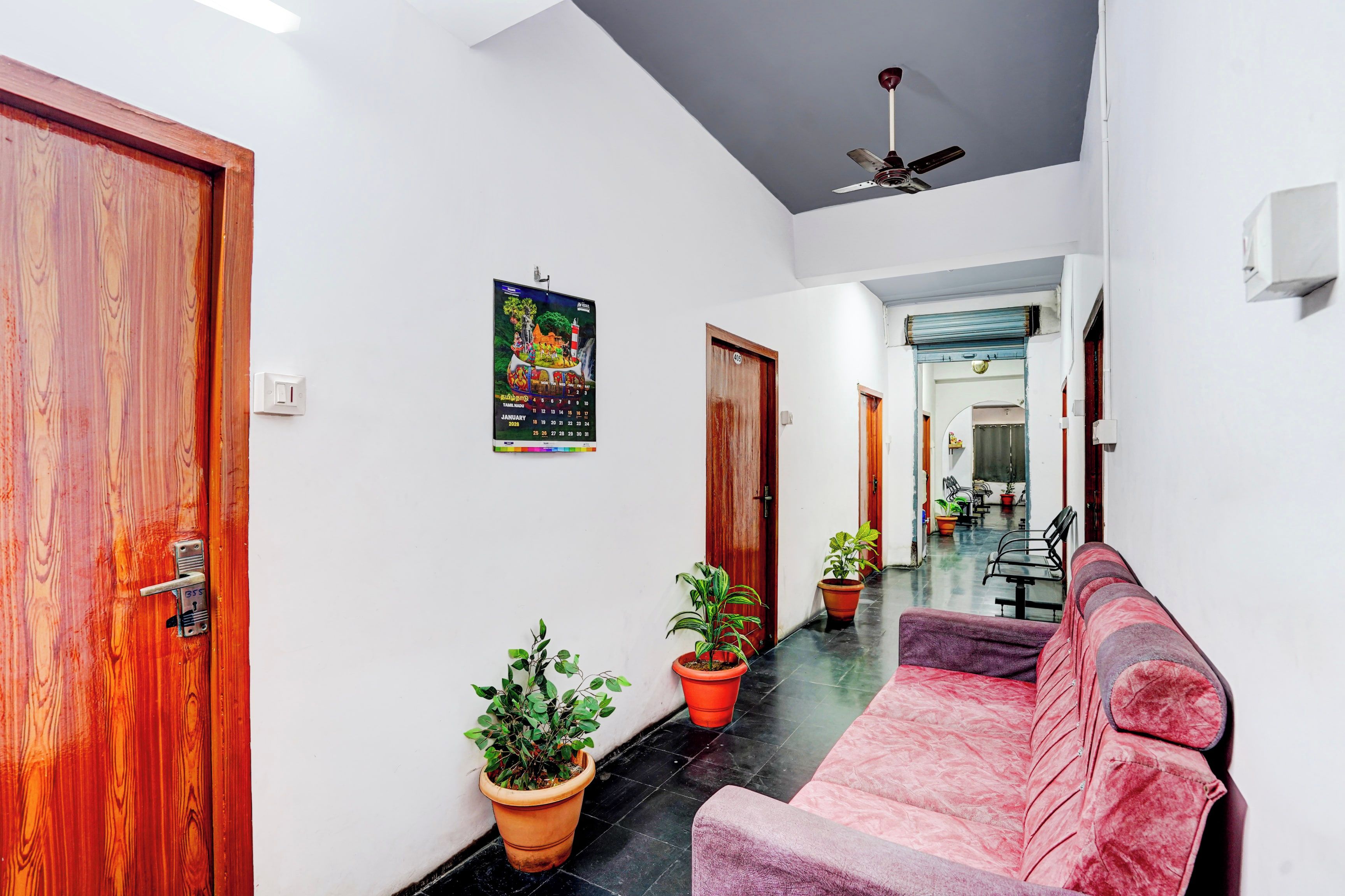 A hallway with a wooden door, a ceiling fan, a calendar, potted plants, and a sofa.