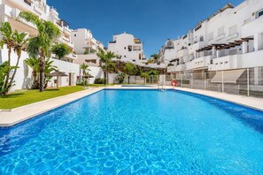 A swimming pool surrounded by white buildings with balconies, a clear blue sky, and some greenery. - VR1B - Golfside Escape Near Estepona (Estepona)