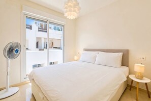 A hotel room with a bed, bedside lamps, a standing fan, and a view through a large window. - VR1B - Golfside Escape Near Estepona (Estepona)