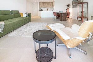 Een moderne woonkamer met een groene bank, een witte leren fauteuil, een ronde zwarte tafel en een keuken op de achtergrond.
