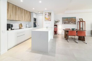 A modern kitchen with white cabinetry, a central island, and a dining area with red chairs. - VV6-2B - Modern 2BR with large seaview balcony (Estepona)