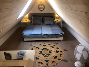 A cozy attic bedroom with a bed, two bedside lamps, a heart-shaped mirror, a woven rug, and a standing fan.