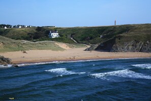 A coastal landscape with a sandy beach, a large white building with multiple windows, and a lighthouse in the distance. - Cottage 'Kêr Didrouz' with Private Terrace, Private Garden and Wi-Fi (Le Palais)