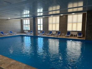 An indoor swimming pool with blue water, surrounded by lounge chairs and large windows. - Felic Home Stylish apartment overlooking the forest (Tsaghkadzor)