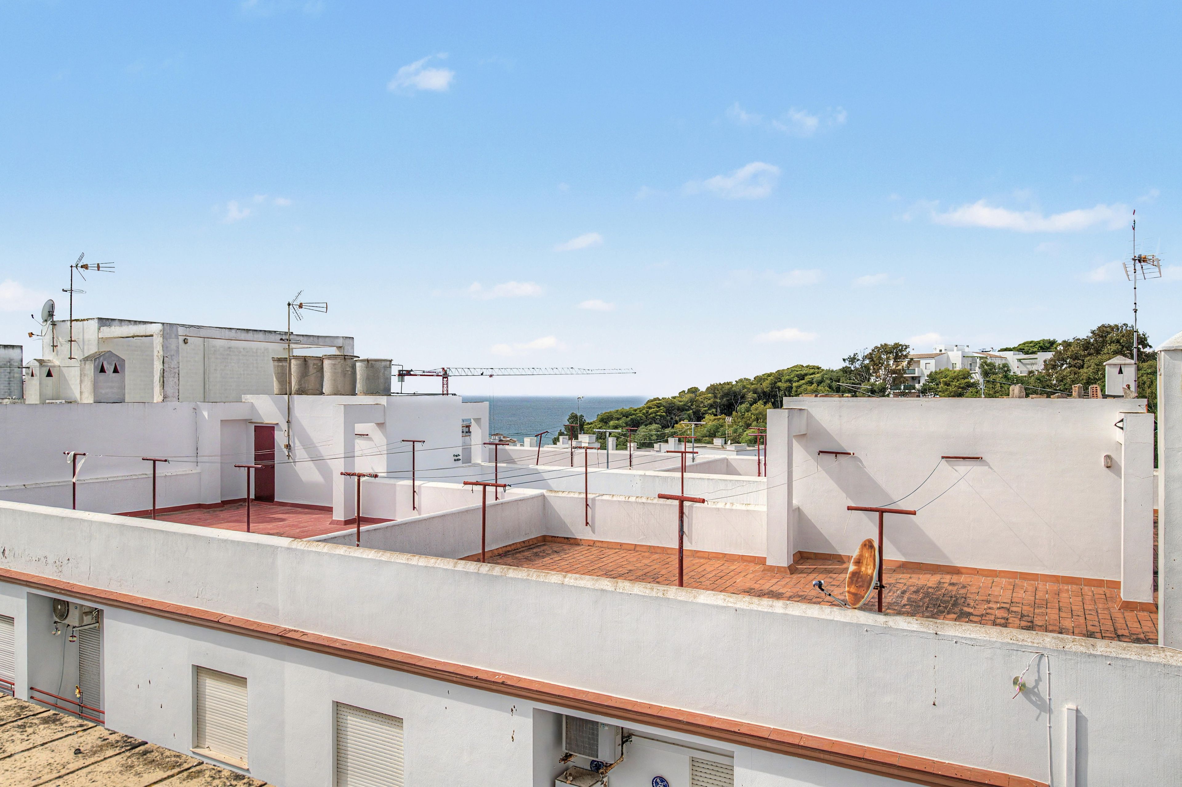 A rooftop terrace with a view of the sea, featuring white walls, red railings, and a brick floor.