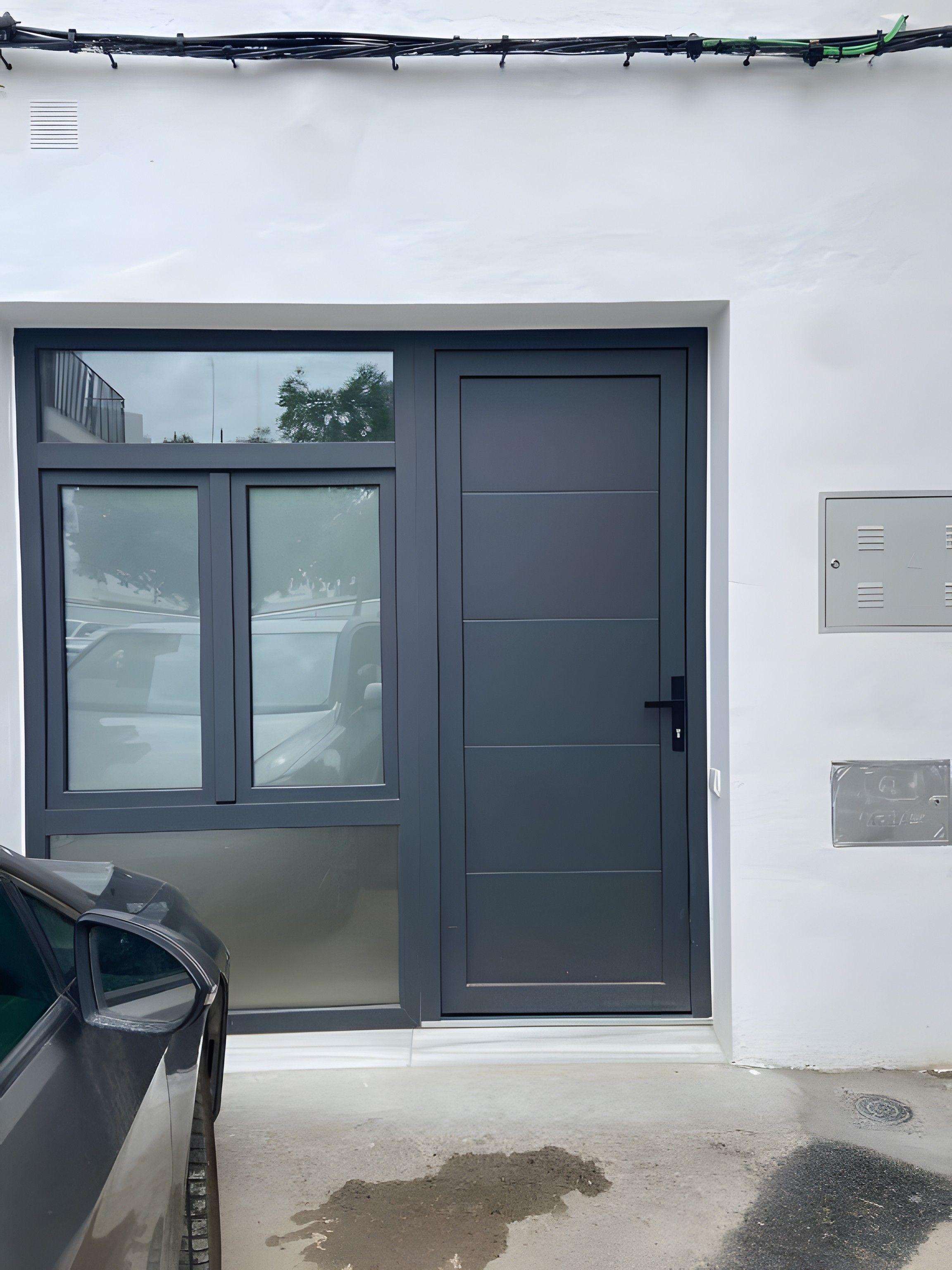 Une entrée de garage avec une porte grise, une voiture garée à l’intérieur et un coffret électrique métallique fixé au mur.
