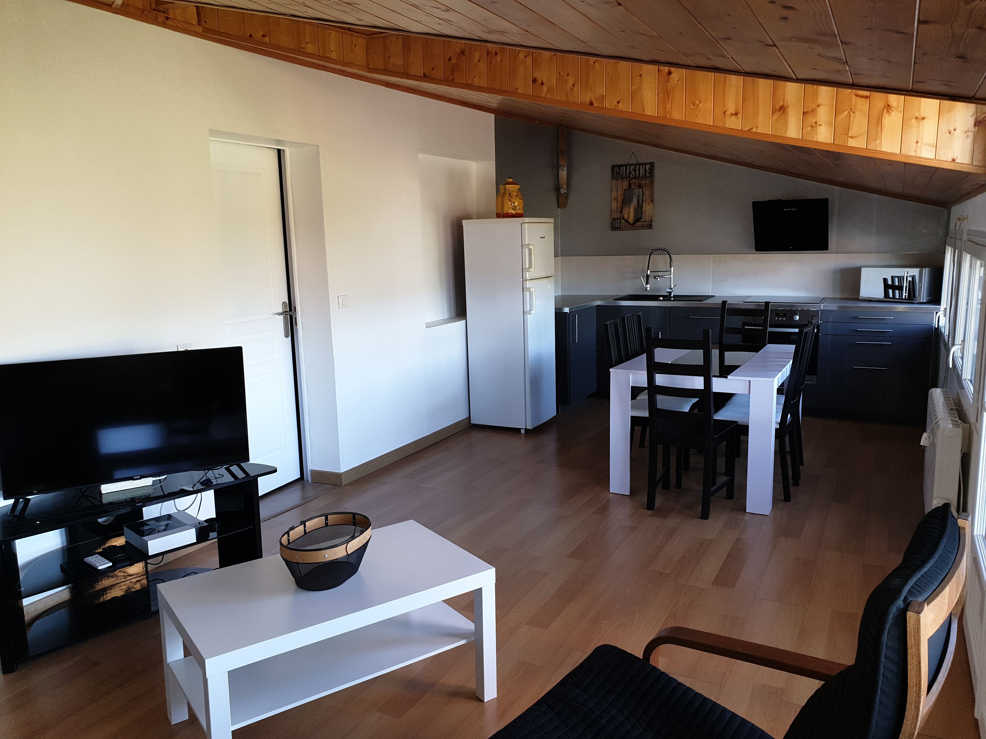 Ein kompakter Wohnraum mit Fernseher, Esstisch, Stühlen und einer Küchenzeile mit Kühlschrank sowie einem Fenster.